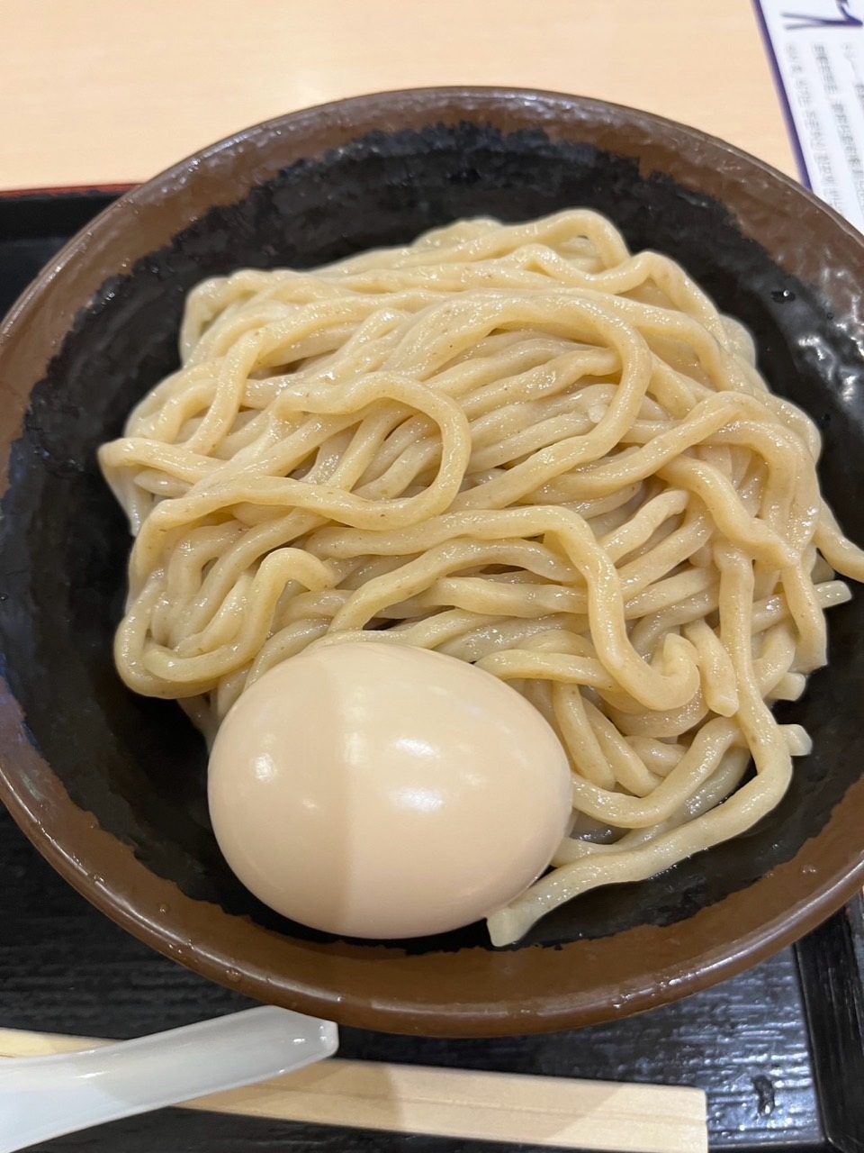 Close-up of thick glossy noodles and a whole egg in a dark-brown bowl.
