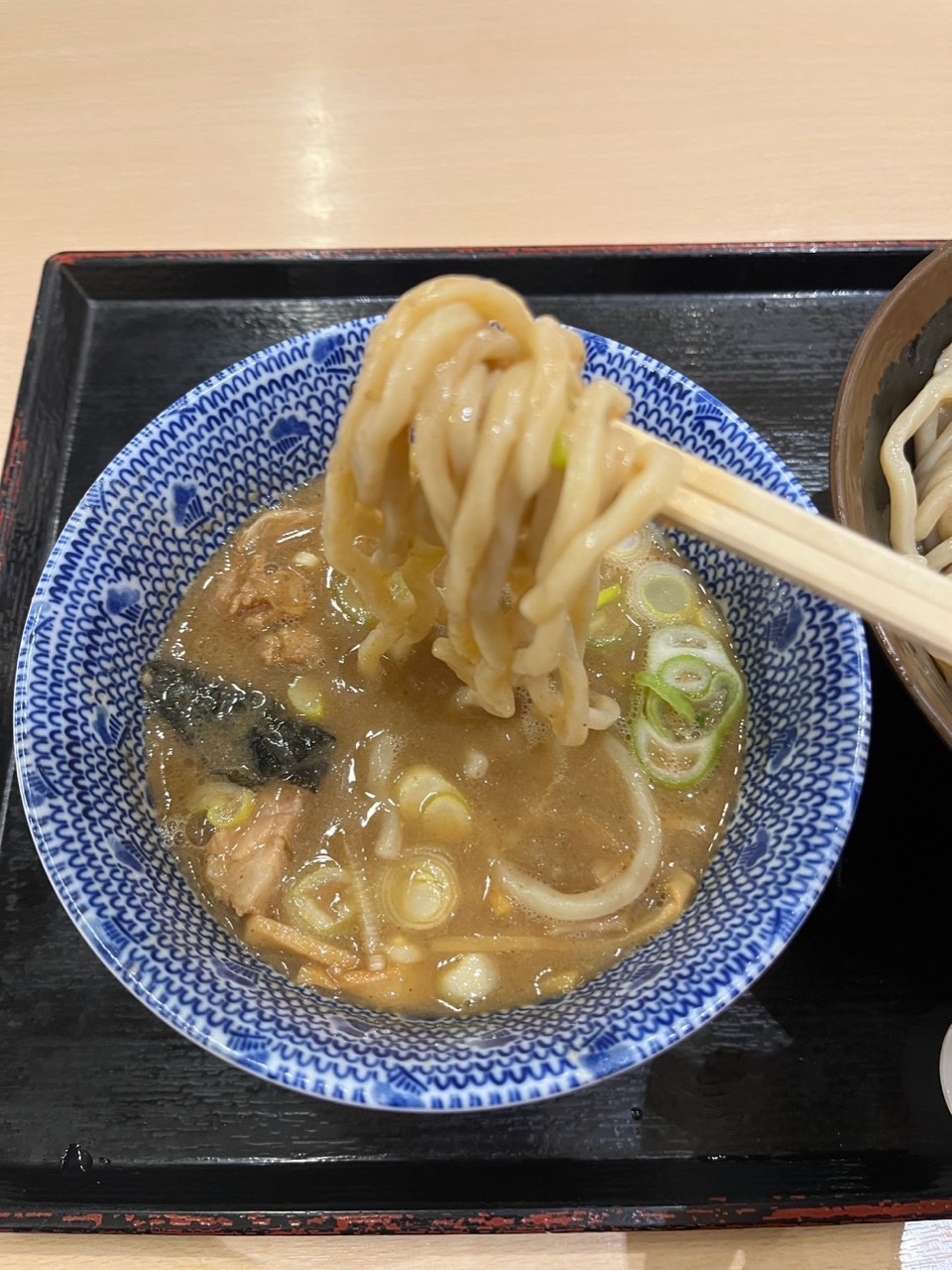 Chopsticks lift thick noodles over tan-brown dipping soup with scallions and nori in a blue-patterned bowl.